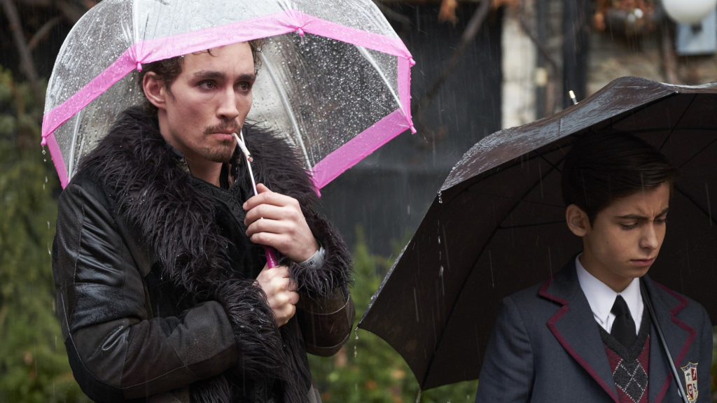 Robert Sheehan sebagai Klaus di The Umbrella Academy memegang payung bening dan merah muda