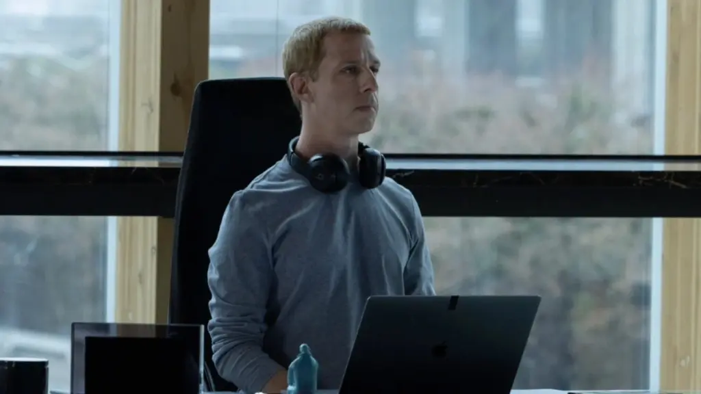 Jeremy Strong berperan sebagai CEO Facebook Mark Zuckerberg yang duduk di mejanya dengan headphone di lehernya.